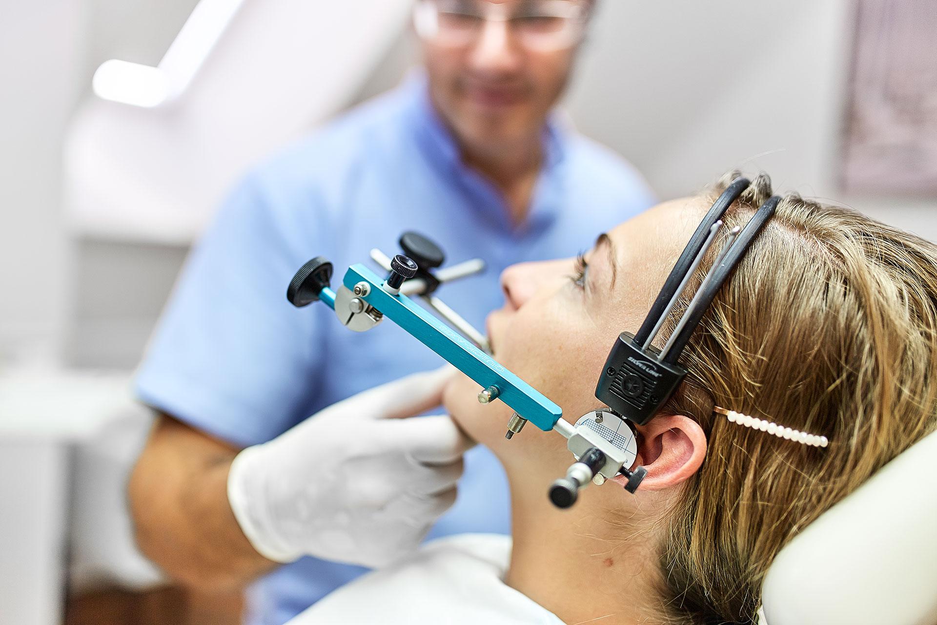 Fogászati rendelőben páciens fején arcregisztrációs eszköz látható, amelyet fogorvos illeszt a fülre és fejre vizsgálat közben, világos háttérrel.